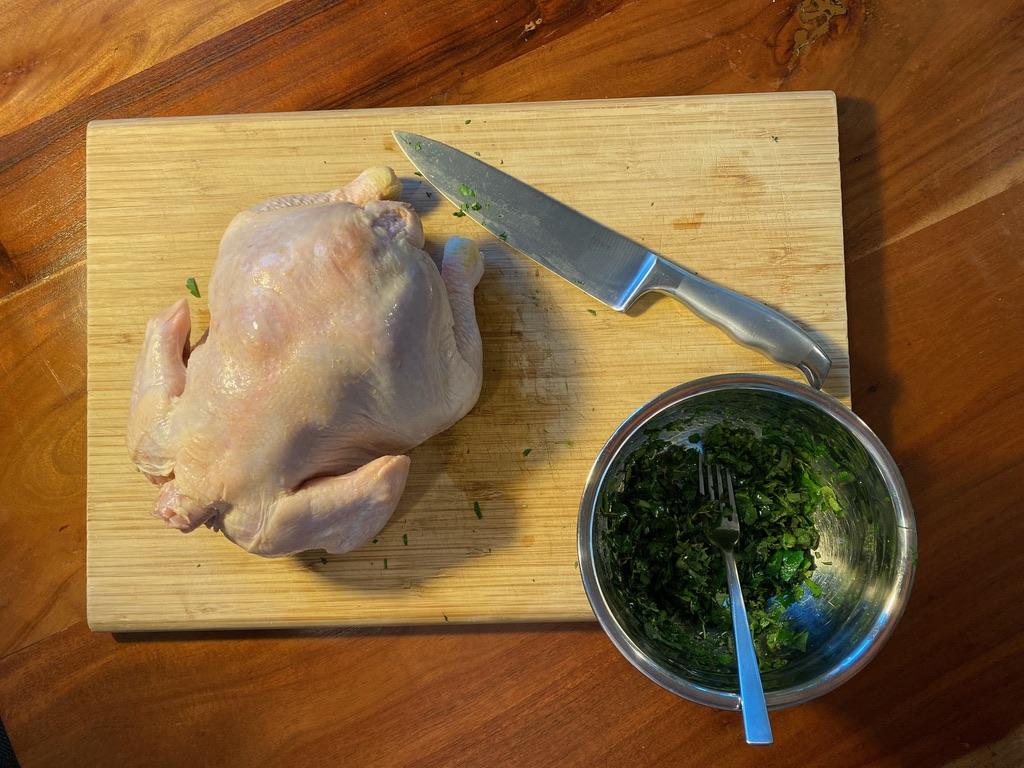 A photo of the top of the previous scene: an uncooked chicken, a mixing bowl (containing herbs), and a knife, all on the same cutting board.