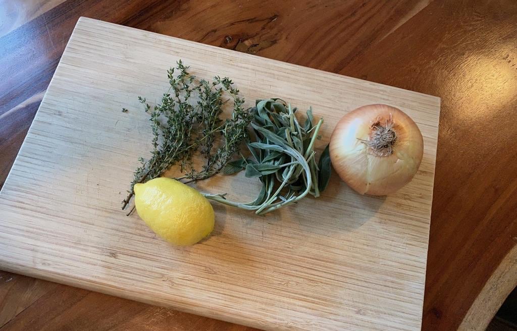 A photo of rosemary, sage, a lemon, and a white onion sitting on a cutting board.